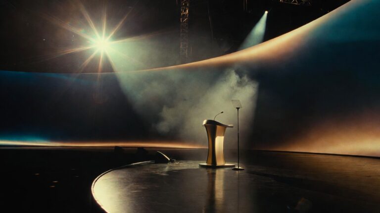 A vast empty premium conference stage moments before the keynote, golden lectern under a single starburst spotlight